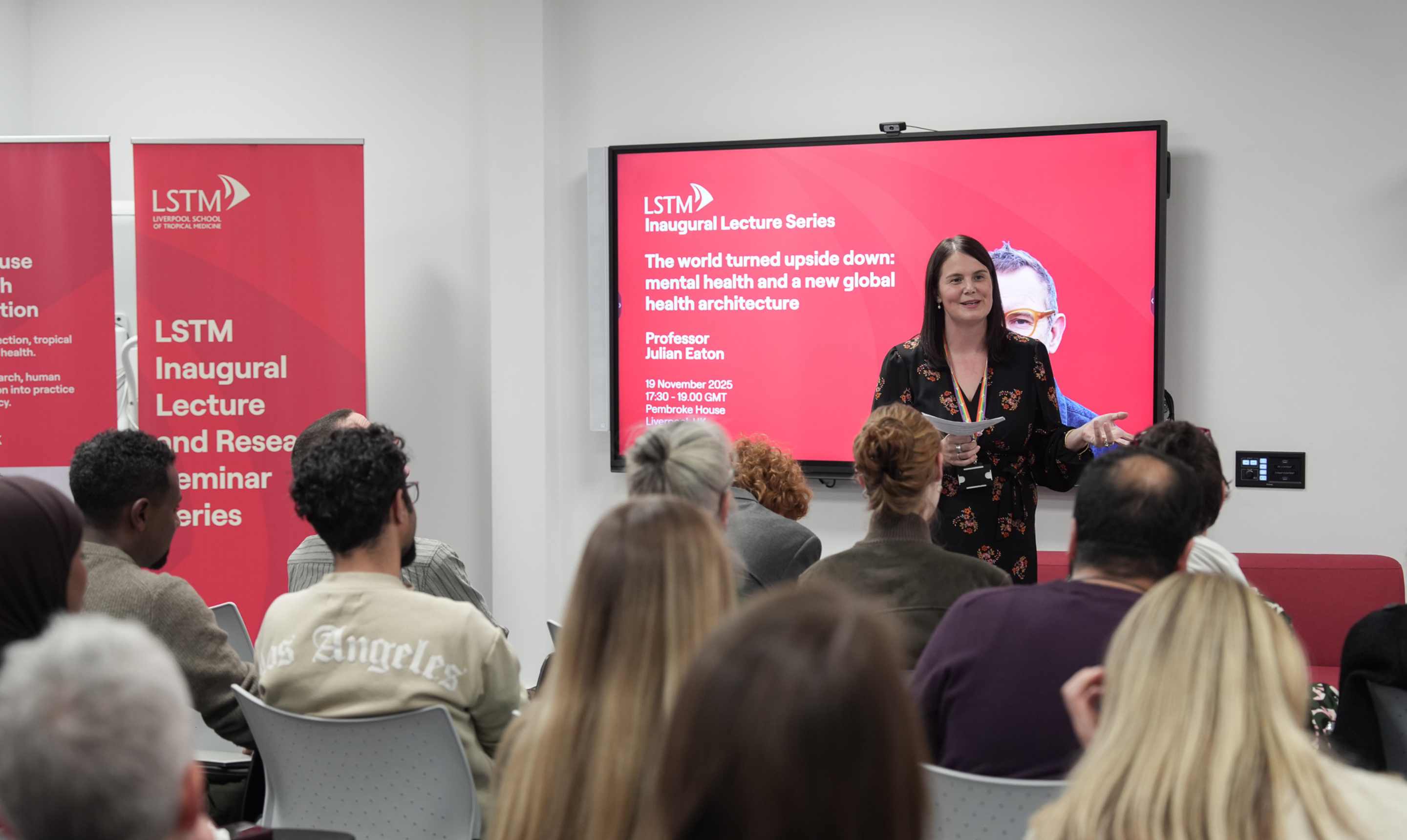 Dr Laura Dean stands at the front of a lecture room speaking to a seated audience, with red LSTM Inaugural Lecture Series banners and a screen behind her displaying details of Professor Julian Eaton’s talk on mental health and a new global health architecture.
