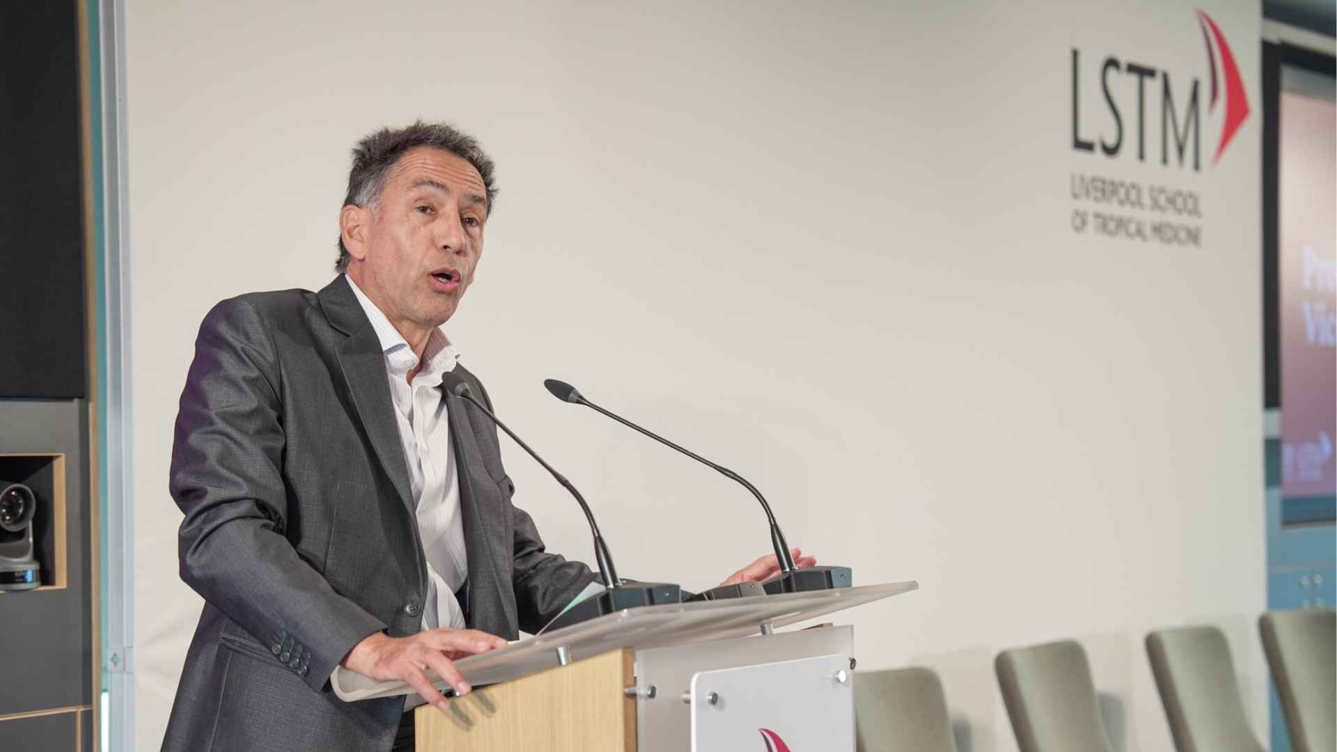 Professor David Lalloo speaks at a lectern with microphones in an LSTM lecture theatre, with the Liverpool School of Tropical Medicine logo and a row of empty chairs visible in the background.