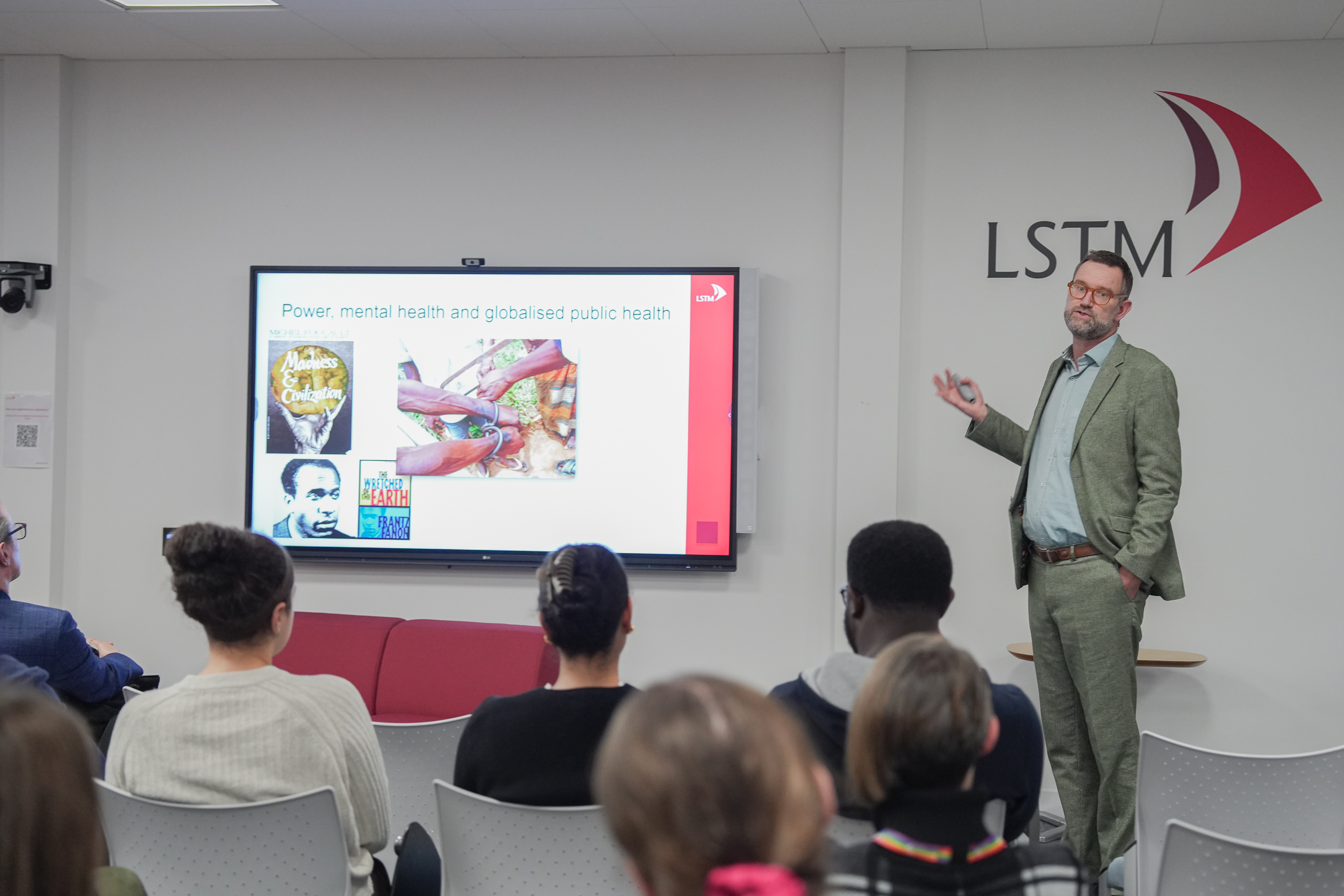 Professor Julian Eaton delivers a lecture at LSTM, standing beside a large screen showing a slide titled “Power, mental health and globalised public health” as an audience sit in rows facing him.