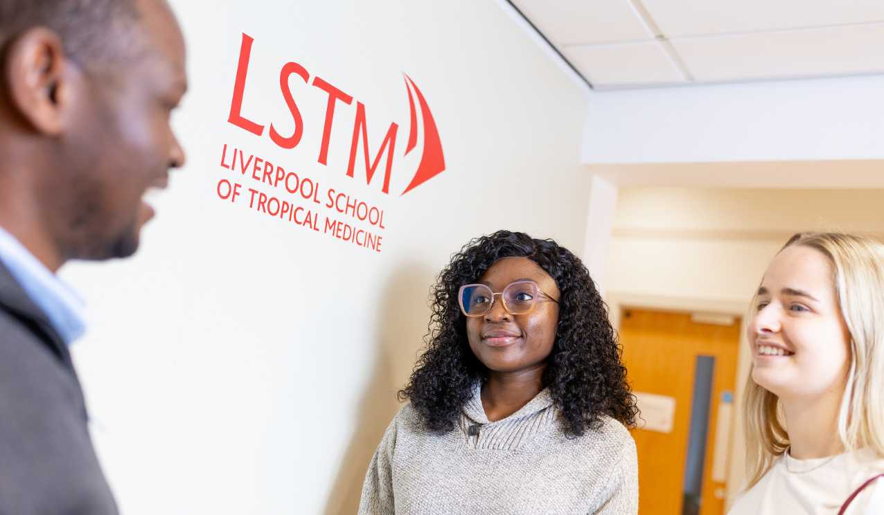 Three students talking in a bright corridor at Liverpool School of Tropical Medicine, with the red LSTM logo visible on the wall behind them.