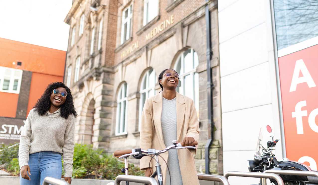 Two students laughing outside Liverpool School of Tropical Medicine, one walking with a bicycle in front of the historic Tropical Medicine building.
