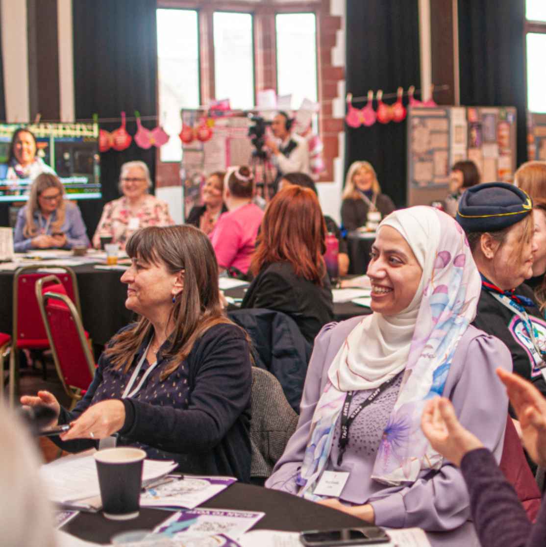 A diverse group of people sit around round tables at a busy conference or workshop, smiling and clapping. In the centre, a woman in a light purple top and patterned hijab laughs with others, while posters, hanging decorations and screens create a colourful, lively backdrop.