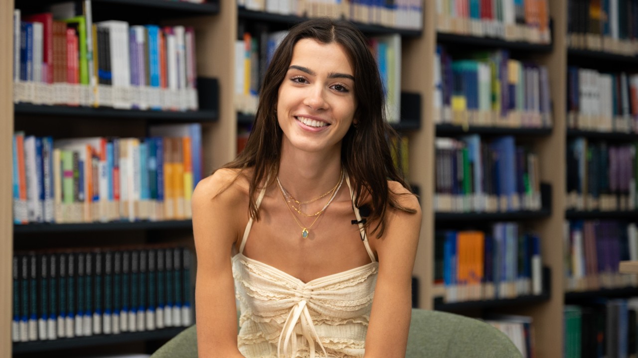 Smiling young woman seated in a library, wearing a cream strappy top and layered necklaces with a clip-on microphone on her strap, with rows of colourful bookshelves softly out of focus behind her.