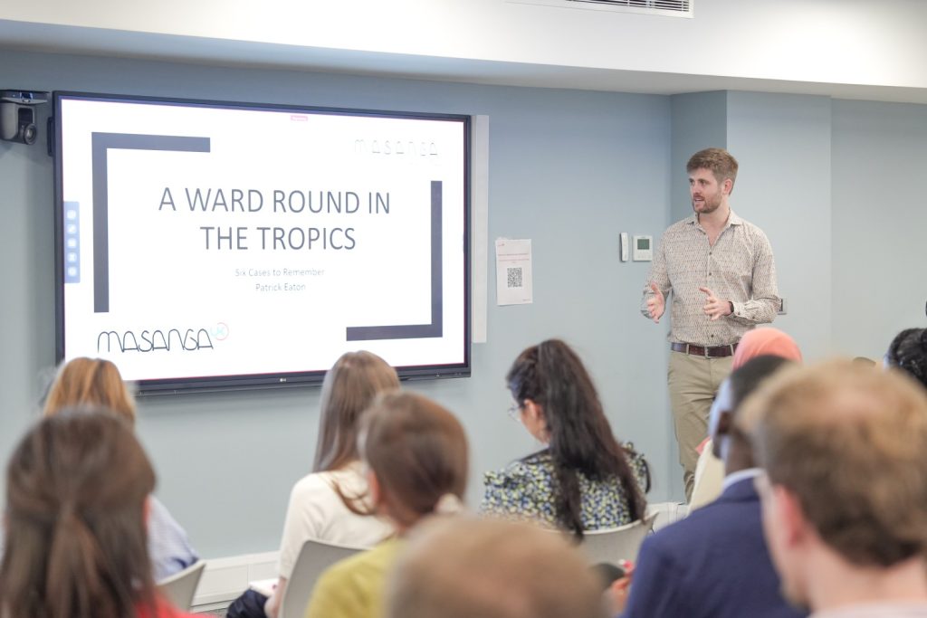 Speaker presenting to a seated audience in a classroom, standing beside a large screen showing a slide titled “A Ward Round in the Tropics – Six Cases to Remember – Patrick Eaton” with the Masanga UK logo.