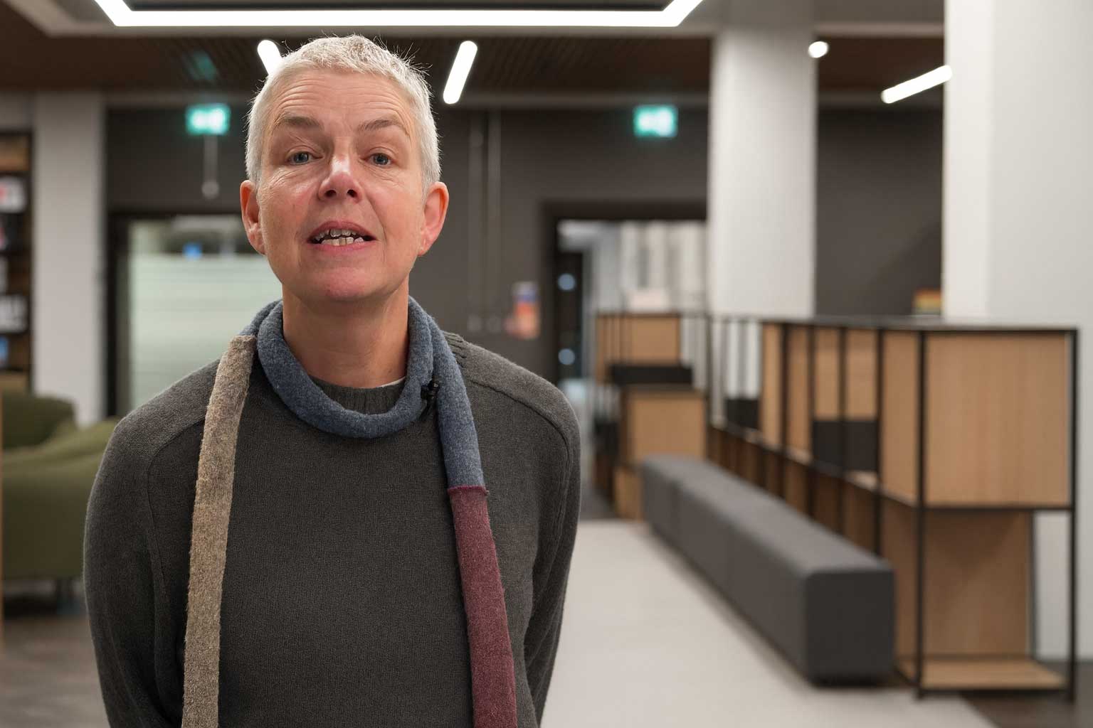 A person with short grey hair, wearing a dark jumper and a long multicoloured scarf, stands in a modern, open-plan interior with soft seating, wooden shelving and bright ceiling lights out of focus in the background.