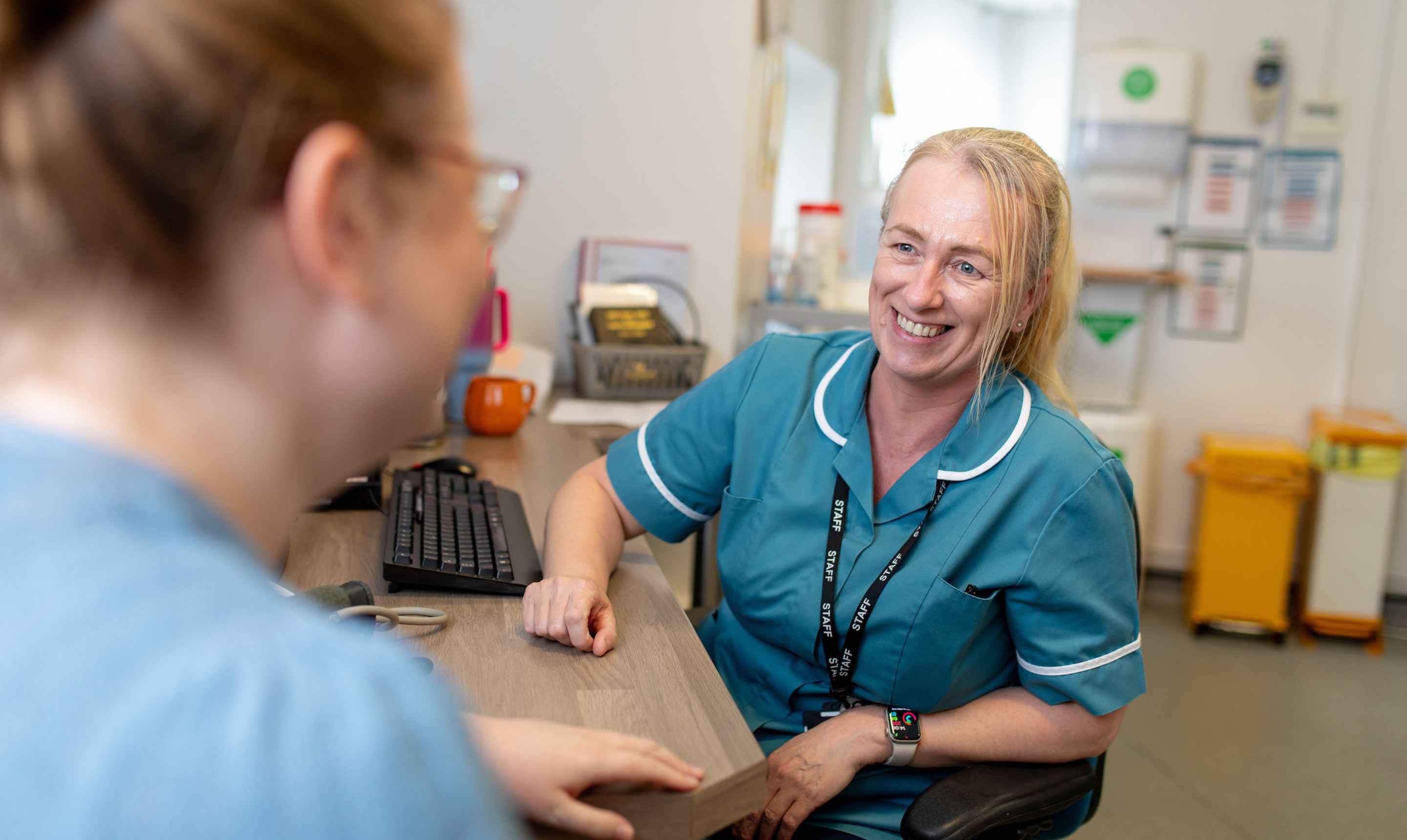 Nurse in teal uniform smiling and talking with a patient across a clinic desk, with a computer and medical equipment visible in the background.