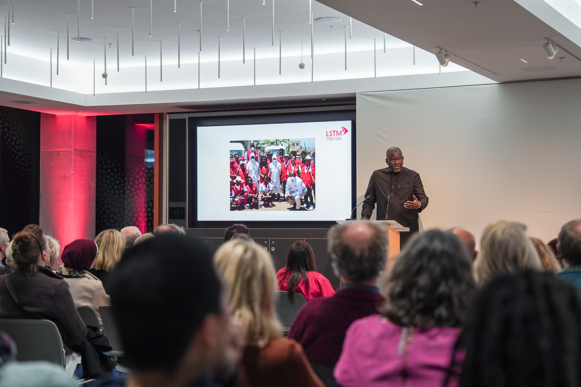 LSTM Chancellor Elhadj As Sy stands at a lectern addressing an audience, with a large screen behind him showing a group photo and the LSTM logo in a modern lecture theatre lit with red accent lighting.