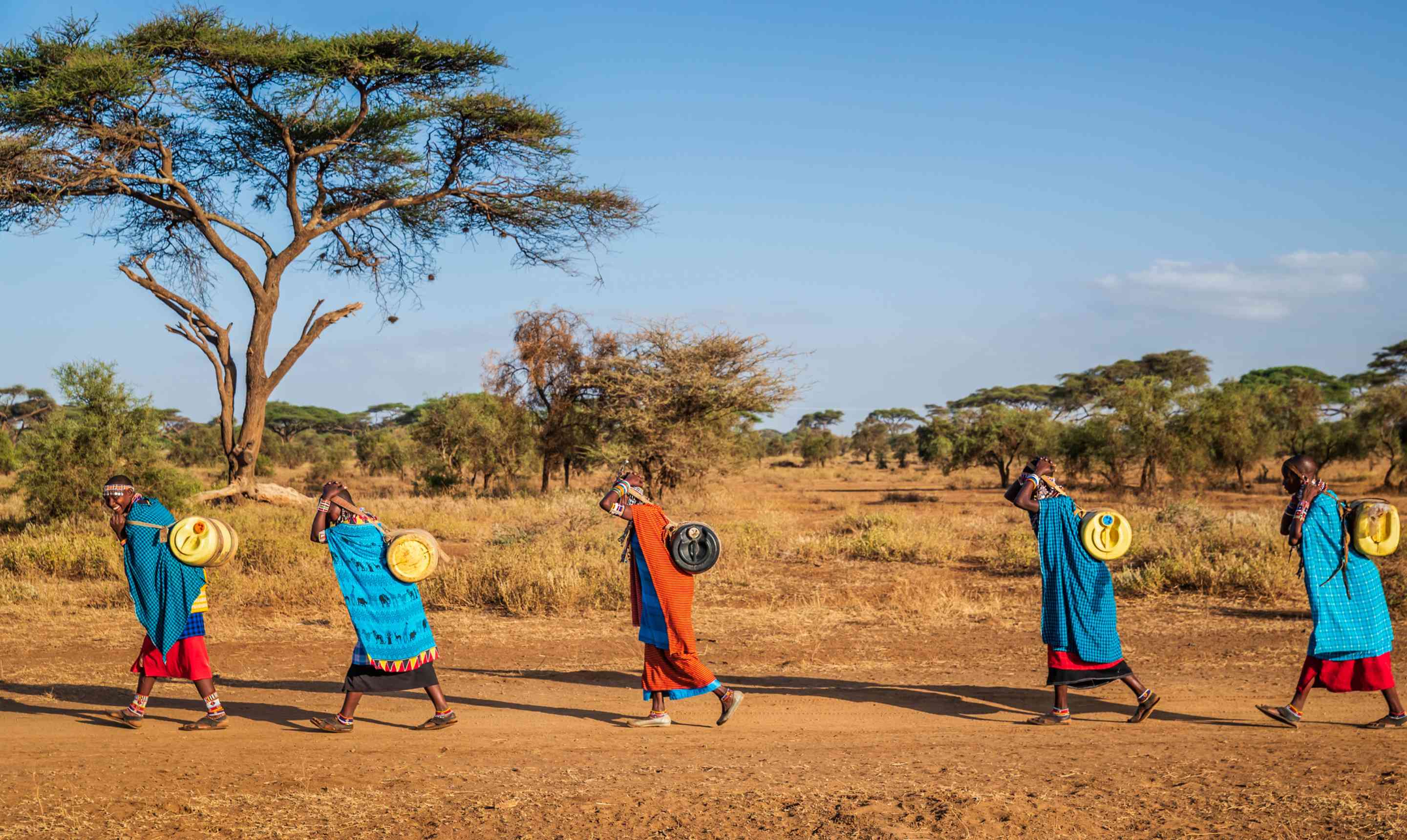 Five people walk in single file across a dry savannah landscape, carrying yellow water containers on their backs beneath a wide blue sky.