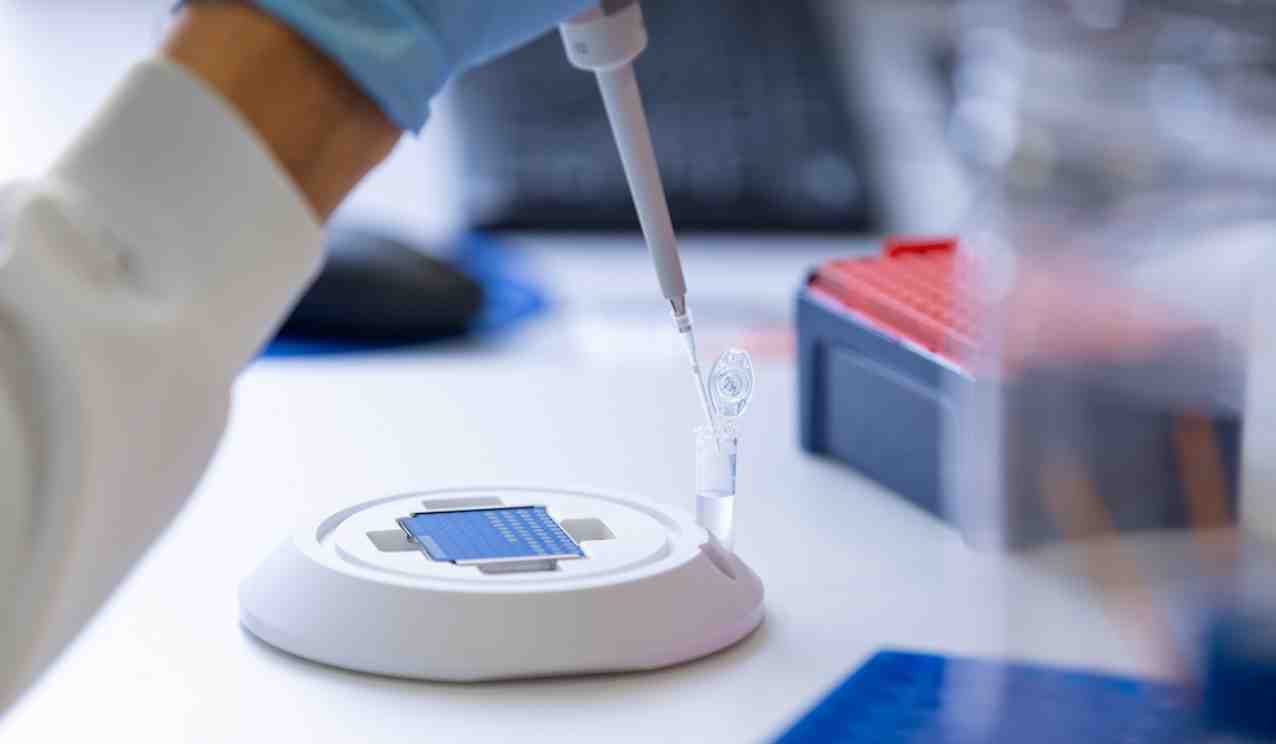 Close-up of a gloved hand using a pipette to dispense liquid into a small tube beside a laboratory testing device.