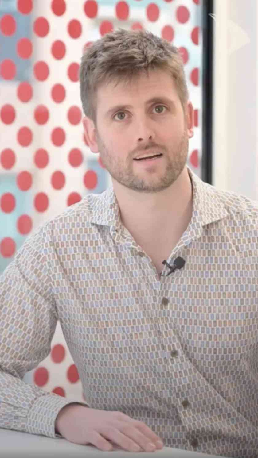 A man with short brown hair and light stubble, wearing a patterned button-up shirt and a clip-on microphone, looks towards the camera while seated indoors, with a red polka-dot wall in the background.