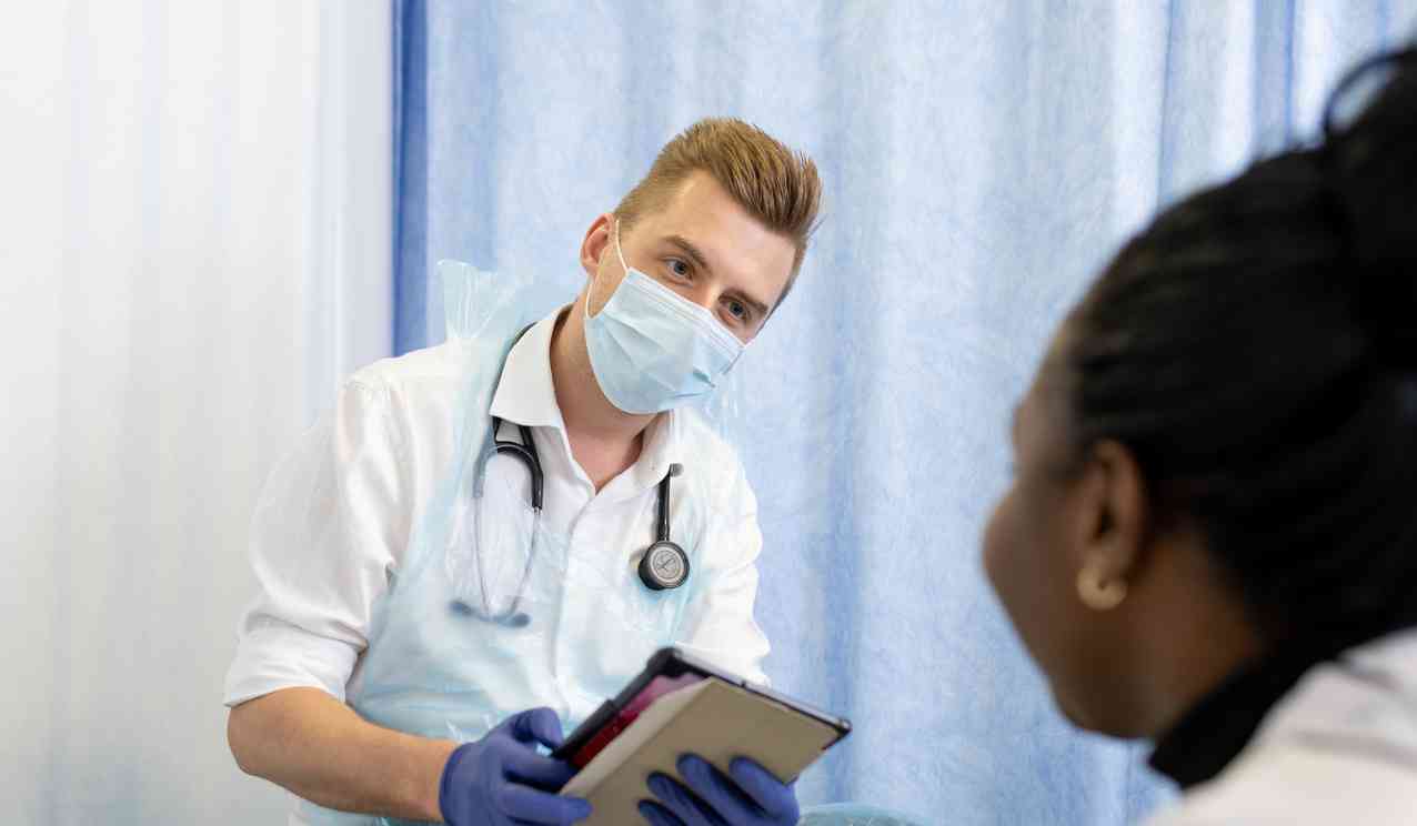Masked clinician with a stethoscope holds a tablet and speaks to a patient during a consultation, with a blue privacy curtain in the background.