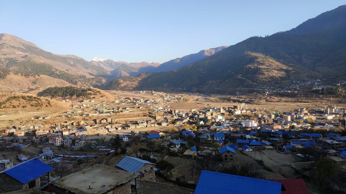Wide view of a small town spread across a valley, with many blue-roofed buildings and dry hillsides, backed by forested mountains under a clear sky.