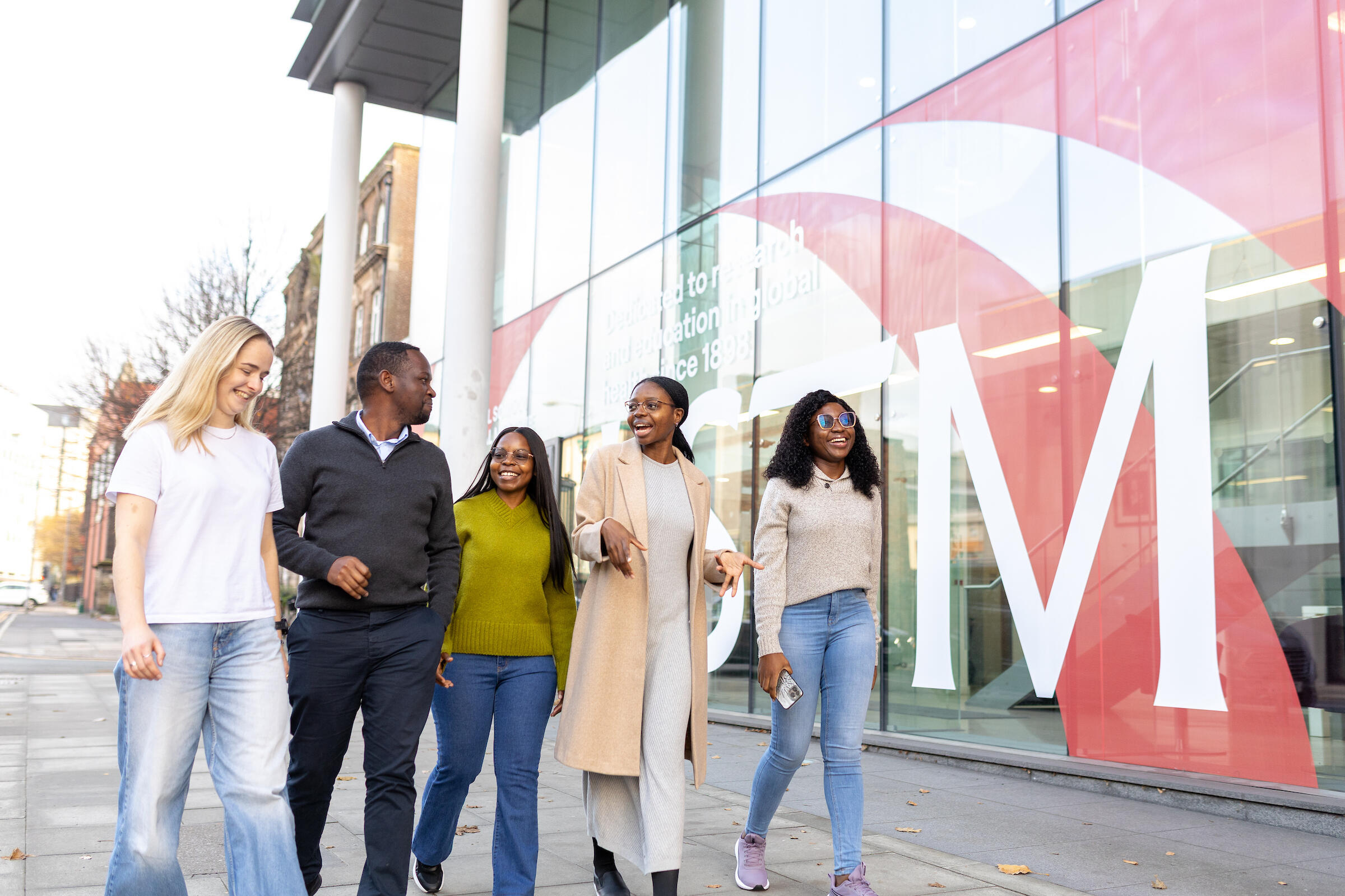 Five people walk and chat along a pavement outside a modern glass-fronted building with large “LSTM” lettering on the window.