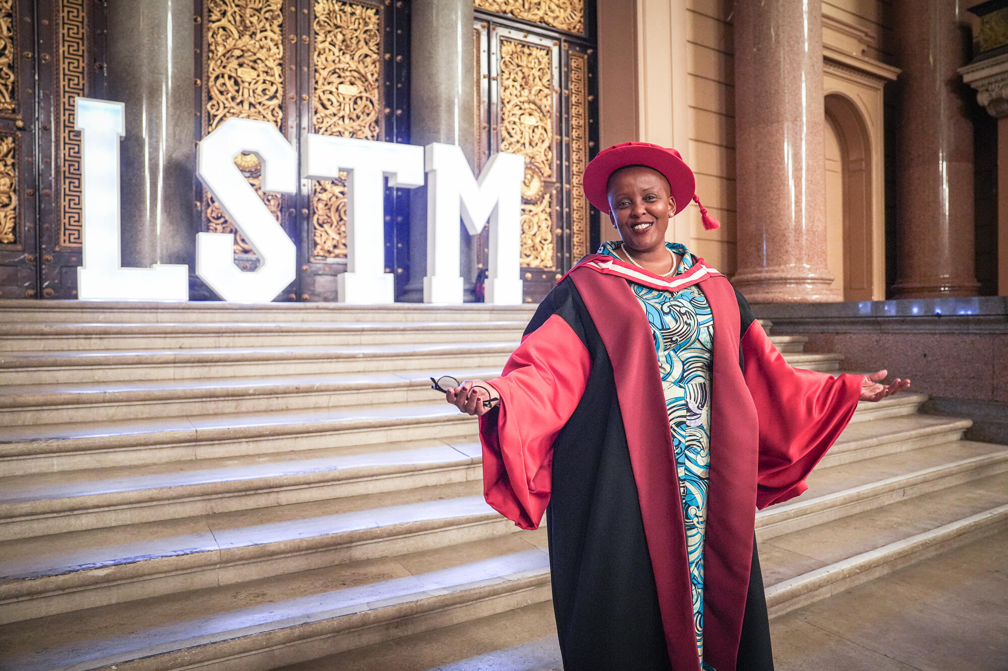 Smiling graduate in red and black academic robes and hat stands on steps inside an ornate hall, with large illuminated “LSTM” letters behind.