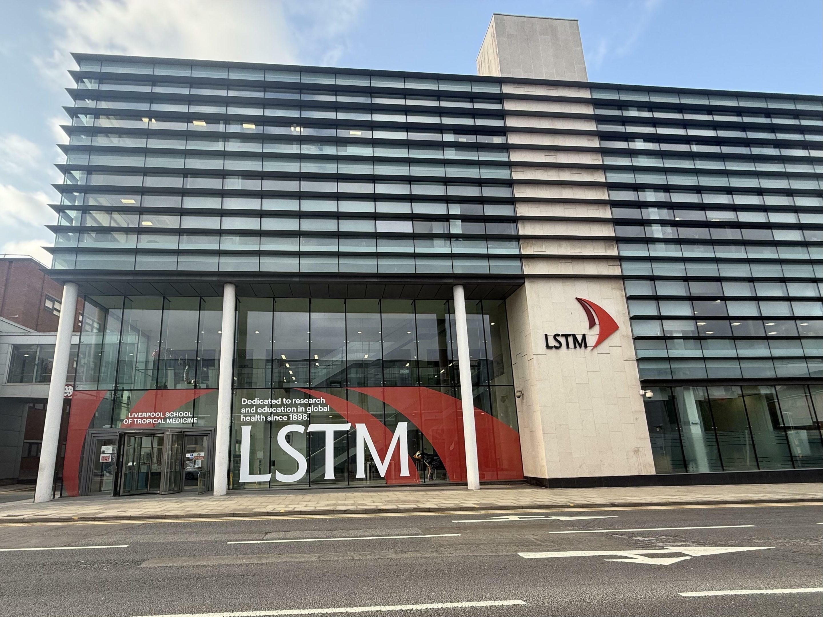 Exterior of a modern glass-fronted Liverpool School of Tropical Medicine building, with large “LSTM” branding and logo on the façade beside a quiet street.