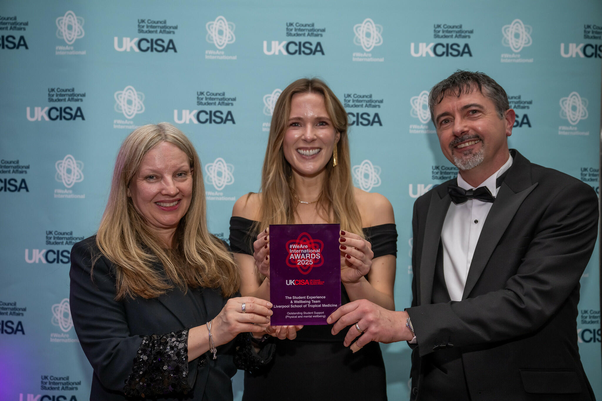 Three people pose in front of a UKCISA backdrop, smiling while holding a purple “#WeAreInternational Awards 2025” plaque.