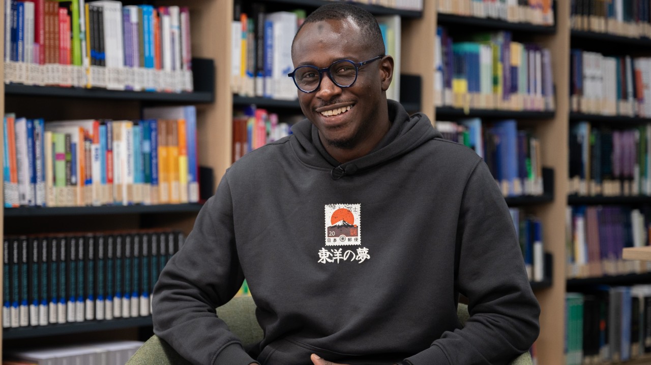 Smiling student wearing glasses and a dark hoodie sits in a chair in front of library bookshelves.
