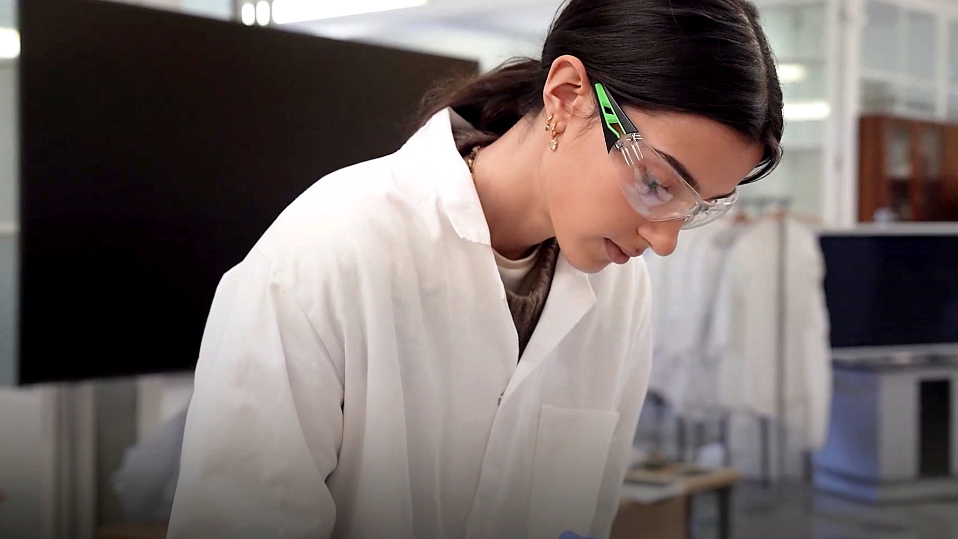 Woman in a white lab coat and clear safety glasses leans over lab work, focused, with a blurred laboratory background.