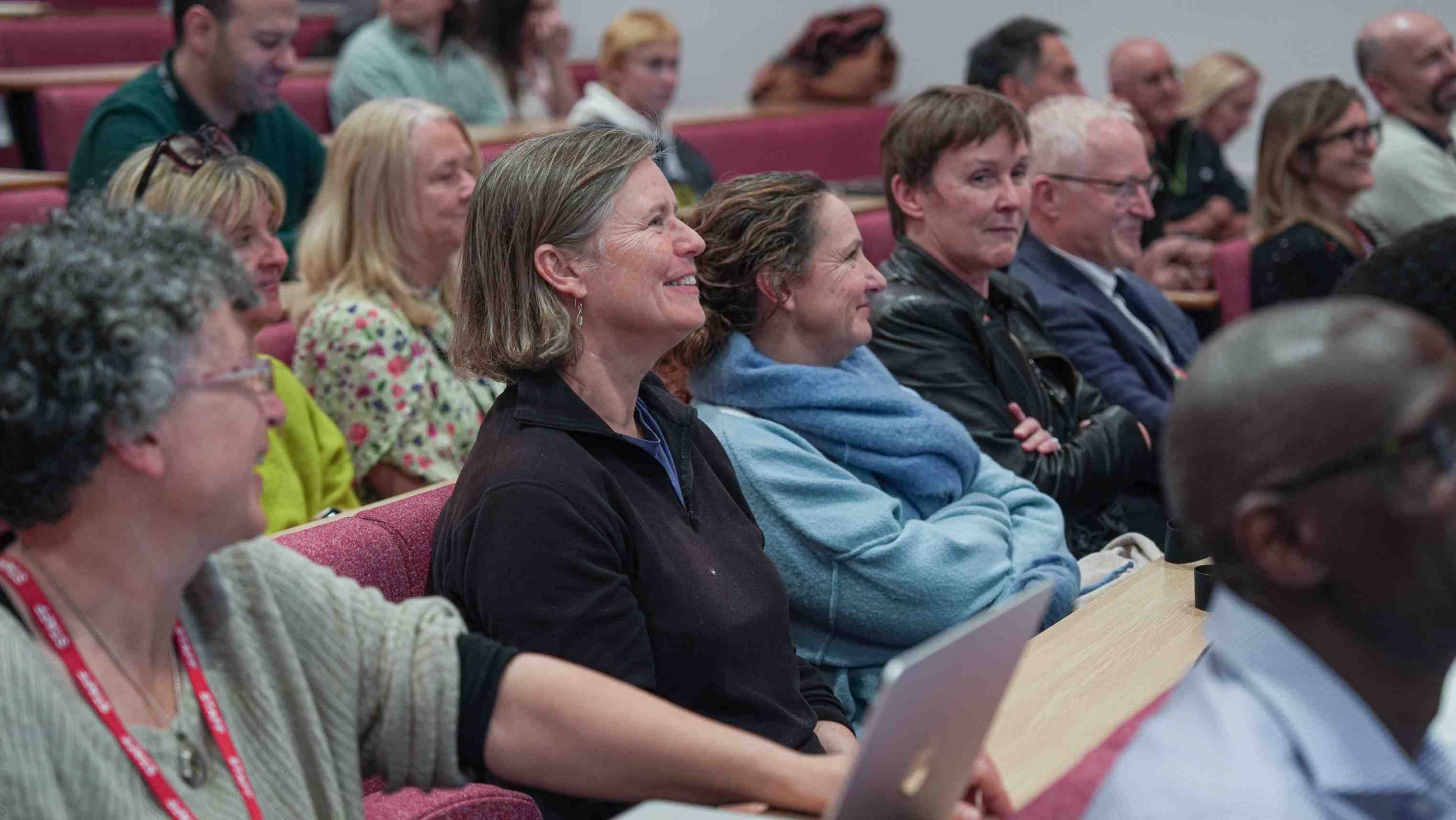 Audience members sit in tiered lecture theatre seating, smiling and listening during a talk.