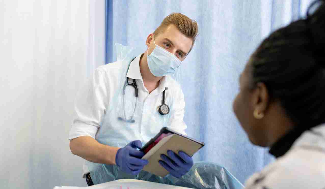 Clinician wearing a face mask and gloves reviews notes on a tablet while speaking with a patient in a clinic room.