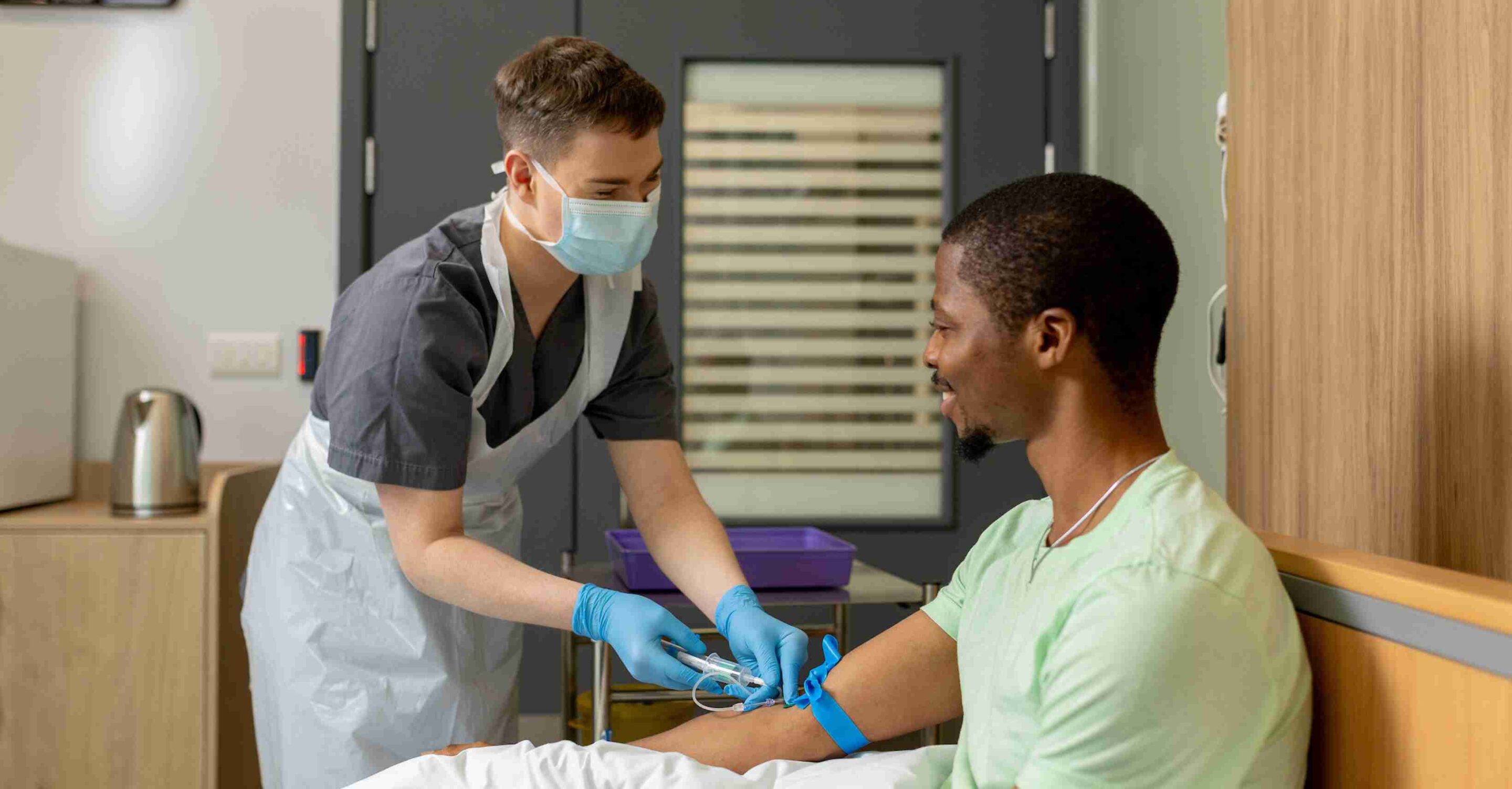 Healthcare worker wearing a mask and gloves prepares to take a blood sample from a seated volunteer’s arm in a clinical room.
