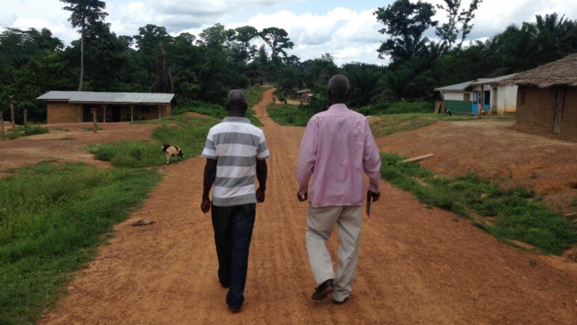 Two men walking away from the camera along a wide, reddish dirt road in a rural village, with scattered houses, greenery, and trees on either side under an overcast sky.