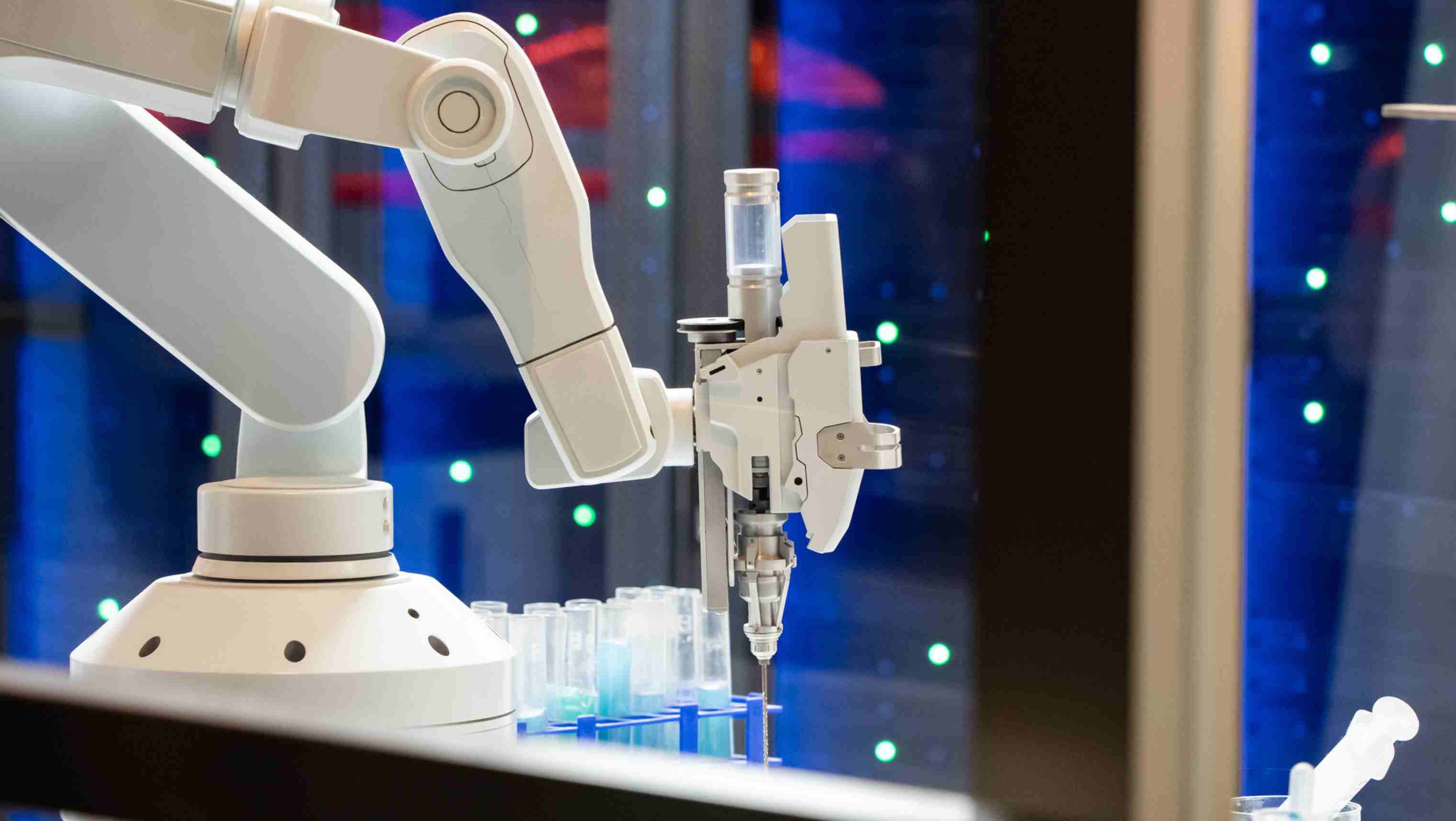 Two white robotic arms in a laboratory setting use syringe-like tools to transfer pink liquid between a small vial and a larger bottle on an automated workstation.