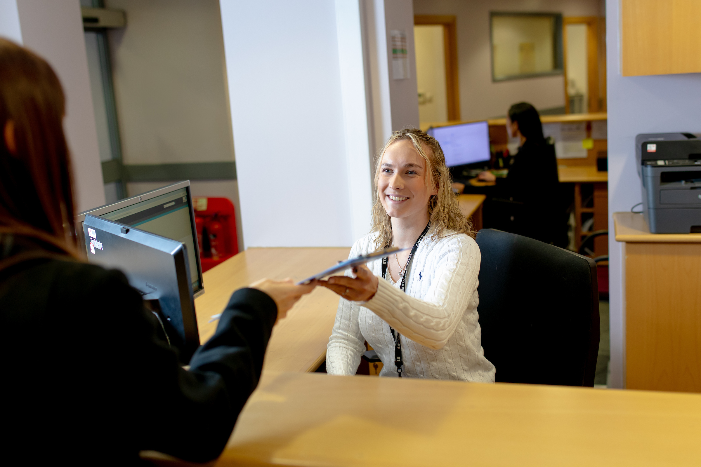 Photo of a member of staff at the Well Travelled Clinic, sitting behind a desk and handing over a form for a visitor to fill out.