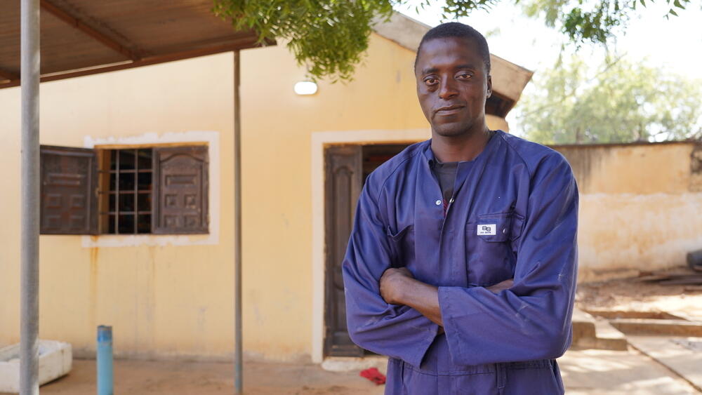 Man in a blue work uniform standing with arms folded outside a clinic building in a sunny courtyard, with trees and a yellow-painted building in the background.