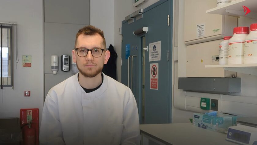 Dr Richard Goodman sits in a laboratory, wearing a white lab coat and glasses, looking directly at the camera. Lab equipment and safety signage are visible in the background.