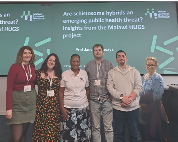 Six researchers stand together in front of a conference slide about the Malawi HUGS project with smiles on their faces.