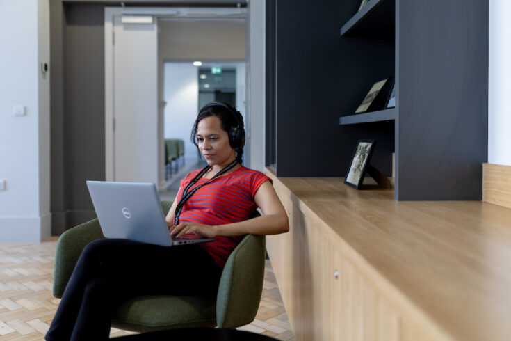 A student sits in a lounge chair using a laptop while wearing headphones in a modern study space with wooden shelving and natural light.