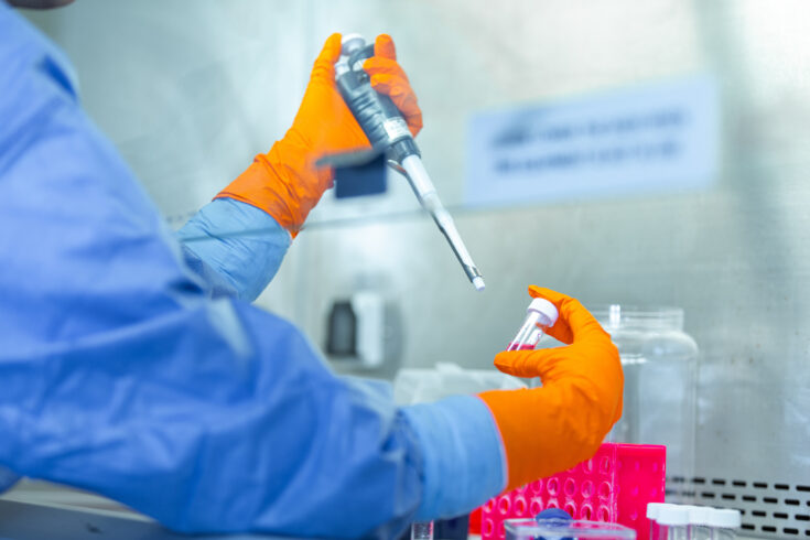 Photo of a person's arms and hands in a lab setting. They are wearing protective equipment, orange gloves and working with test tubes and pipette.