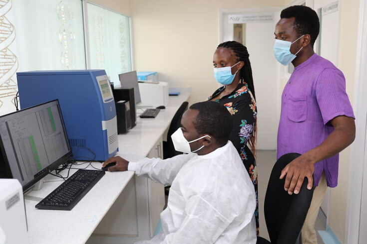 Four people in a laboratory wearing masks; one seated technician works at a computer while three colleagues stand nearby and observe.