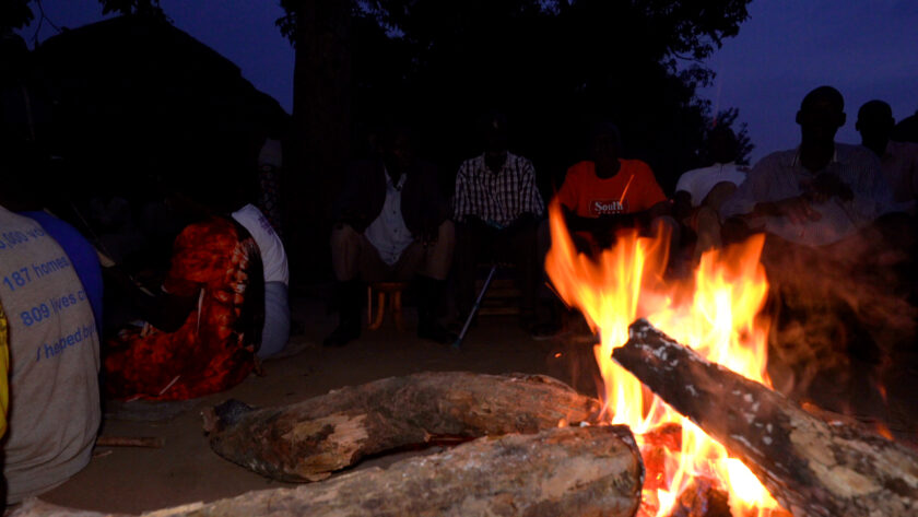 Photo showing a group of people sitting around a campfire at night. The fire is in the foreground to the right of the picture.