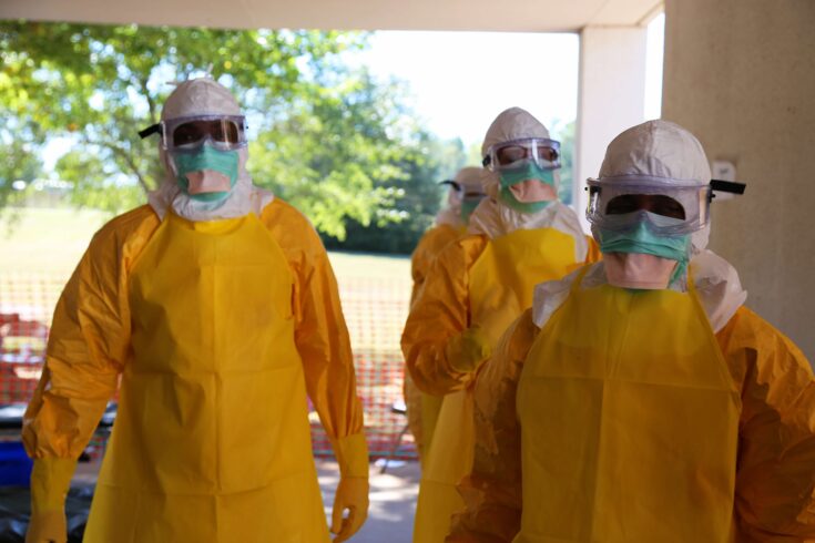 This image was created by Nahid Bhadelia, MD, Assistant Prof. of Medicine in the Section of Infectious Diseases, and the Associate Hospital Epidemiologist at Boston Medical Center. It was captured while the U.S. Centers for Disease Control and Prevention’s (CDC) 2014, Domestic Ebola ETU Training Course for healthcare workers was underway. The program had been designed in order to educate participants who would be deployed, as members of the West African Ebola Response team, as to the proper protocols to be followed when treating Ebola patients. In this particular view, three training participants were displaying the personal protective equipment (PPE) worn by treatment specialists who would be intimately interacting with patients ill with Ebola, which included head and facial coverings, goggles, plastic gloves, a plastic body suite, and plastic apron.