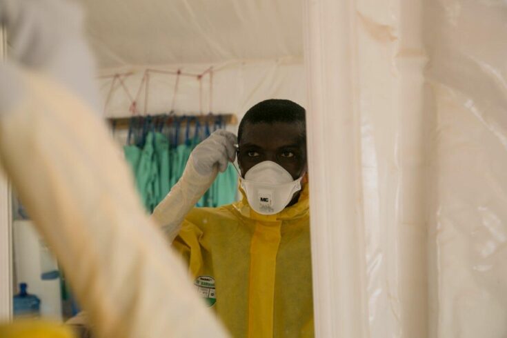 A picture of a man wearing yellow PPE and putting on a face mask.