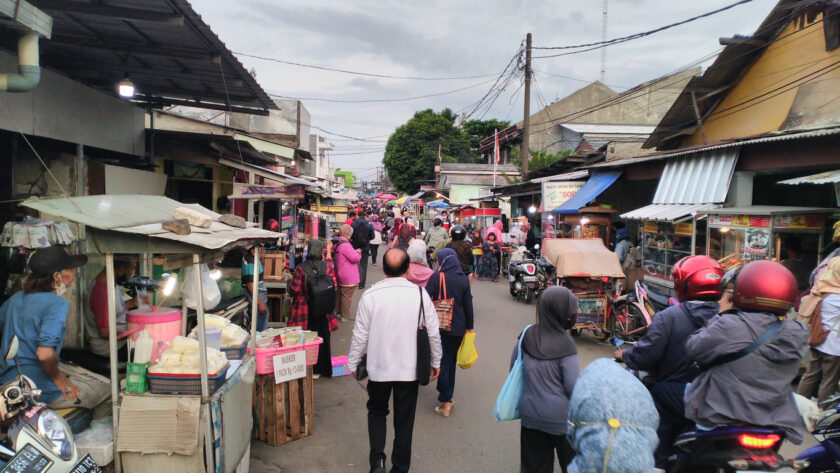 Busy community market in Depok, Indonesia, with pedestrians, vendors and motorcycles along a narrow street.