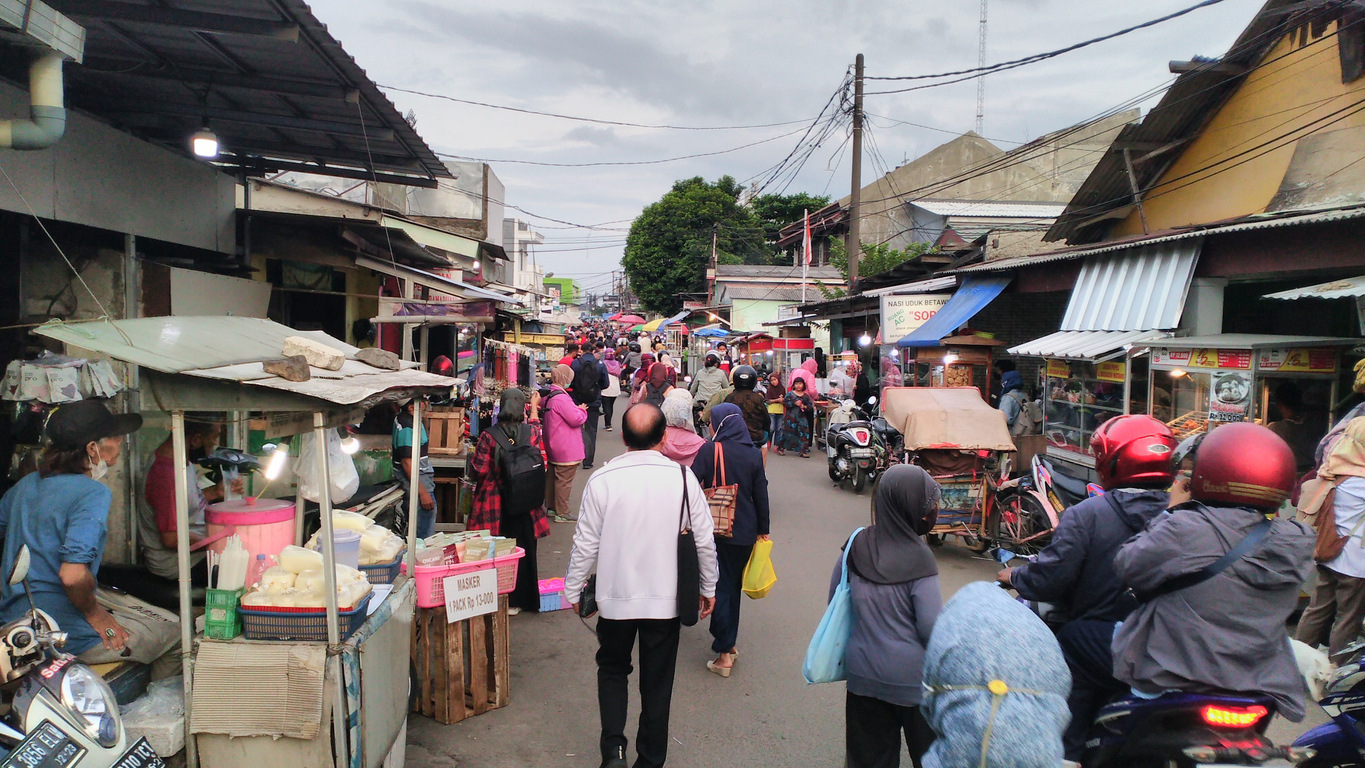 Busy community market in Depok, Indonesia, with pedestrians, vendors and motorcycles along a narrow street.