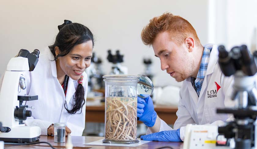Two researchers in lab coats examine a jar filled with worms on a lab bench, with microscopes nearby.