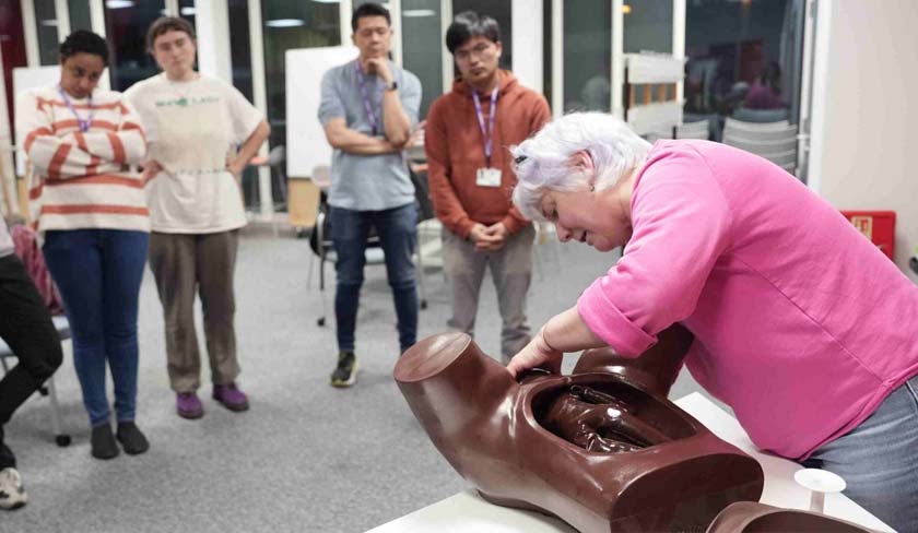 Instructor performing CPR on a large brown training manikin while five students observe in a classroom setting.