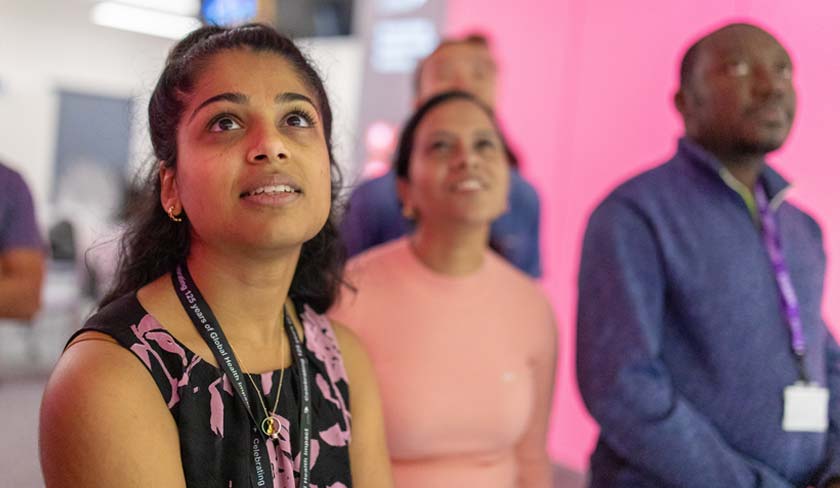 Young woman in the foreground gazes upward at an event, wearing a black and pink patterned top and a lanyard with a badge.